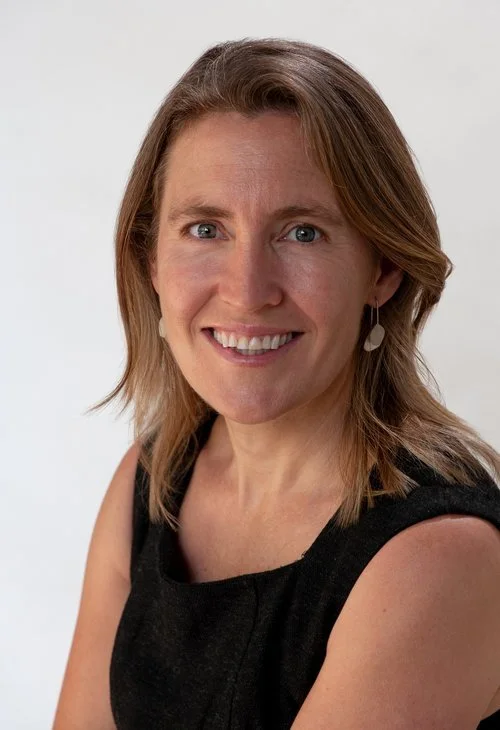 A woman with shoulder-length light brown hair, wearing a black sleeveless top and earrings, smiling at the camera against a plain white background.