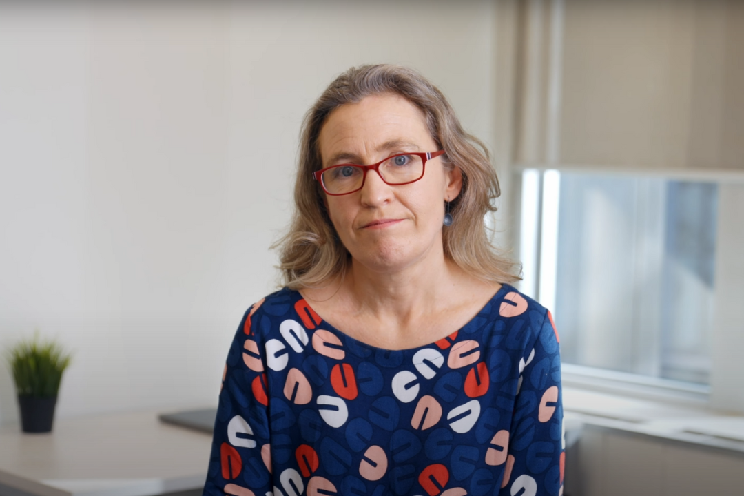 A woman with light brown hair, red glasses, and a blue patterned shirt sitting indoors near a window.