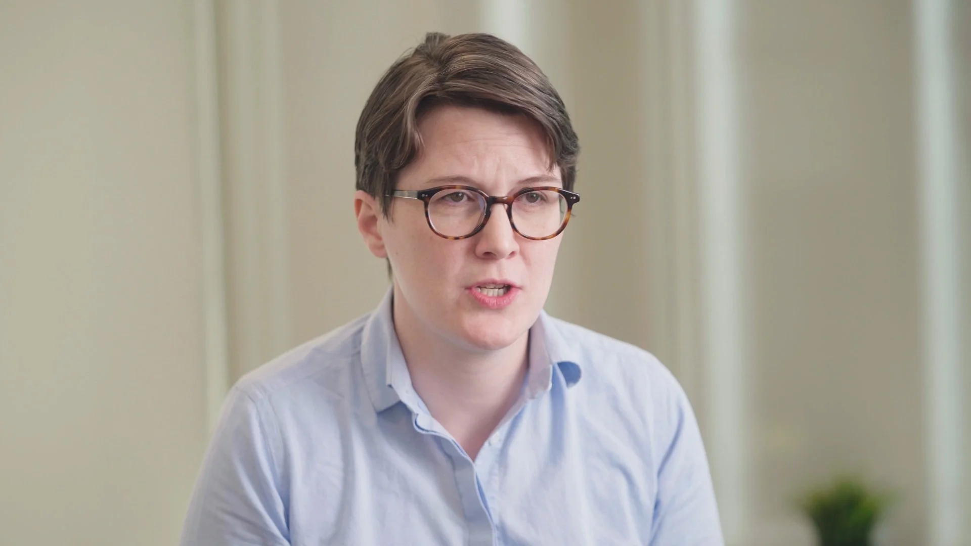 A woman with short brown hair, glasses, and a light blue button-up shirt speaking in an indoor setting with light-colored walls.