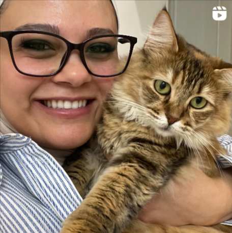 A woman with glasses smiling and holding a tabby cat with green eyes.