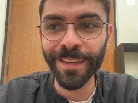 A young man with glasses and a beard smiling in an indoor setting.