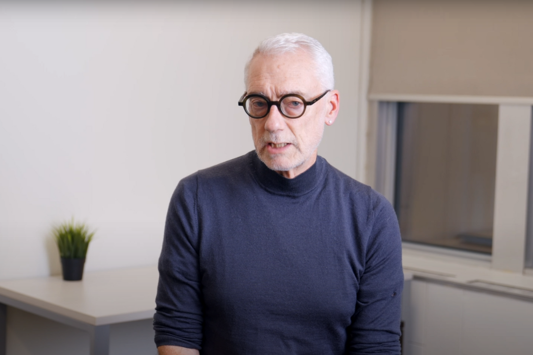 Older man with white hair, glasses, and a black turtleneck speaking in an office setting with a plant on a white desk and a window in the background.