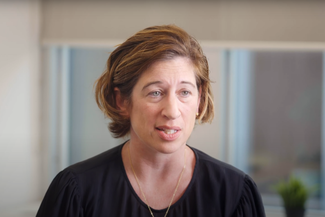A woman with short, wavy, brown hair speaking, wearing a black top and a gold necklace, in a bright interior space.