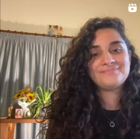 A woman with long, curly dark hair smiling in a room with a beige wall and a wooden shelf behind her, which holds a bouquet of yellow flowers, a framed photo, and a small container.