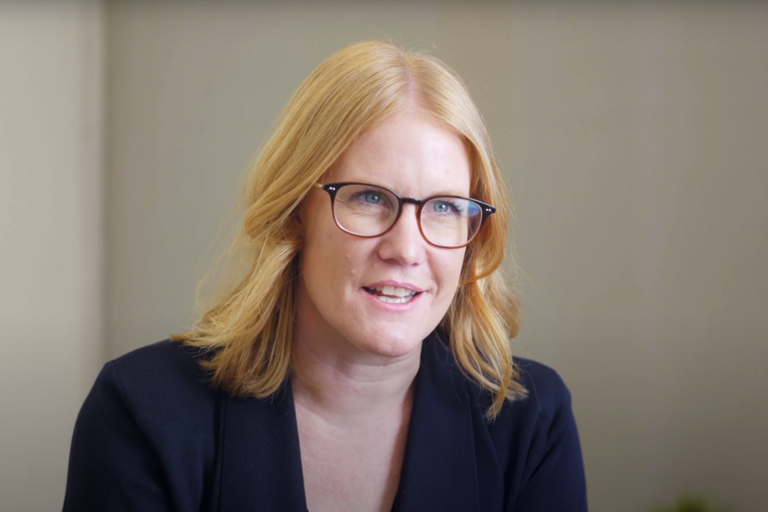 A woman with blonde hair, glasses, and a dark blazer speaking in an indoor setting.