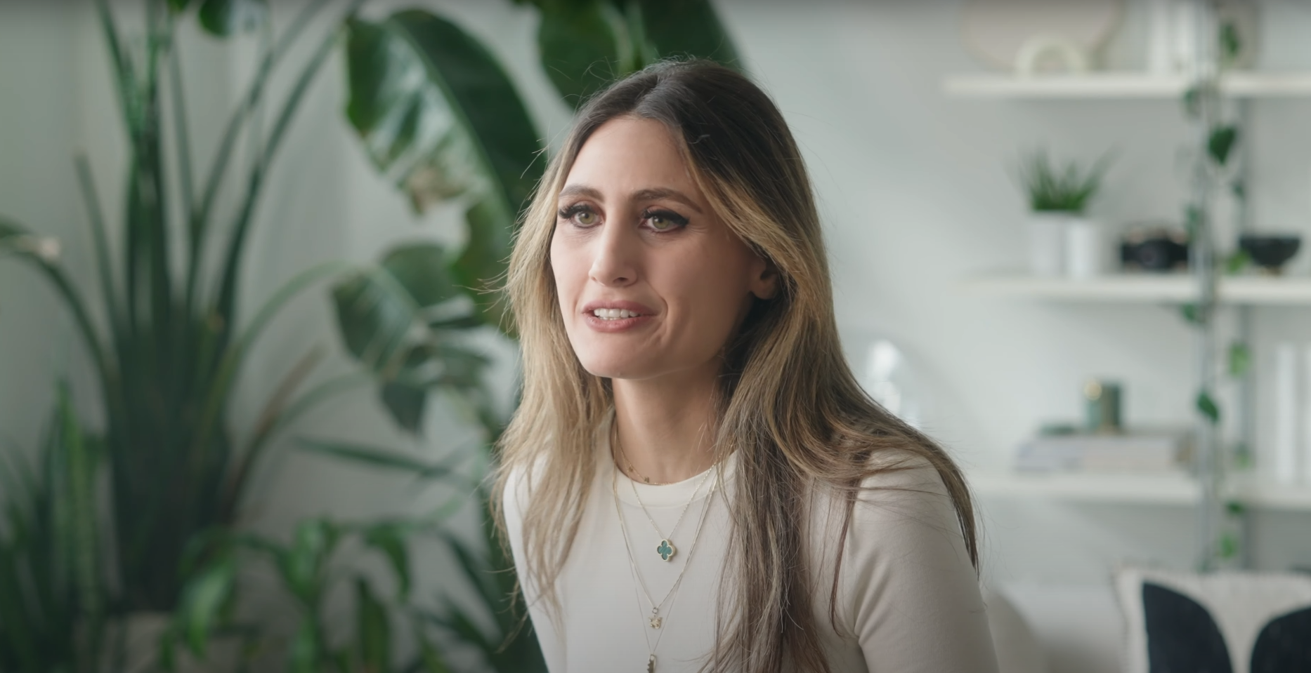 A woman with long brown hair and light makeup, wearing a white top with layered necklaces, sitting in a modern, well-lit room with green plants and white shelves in the background.