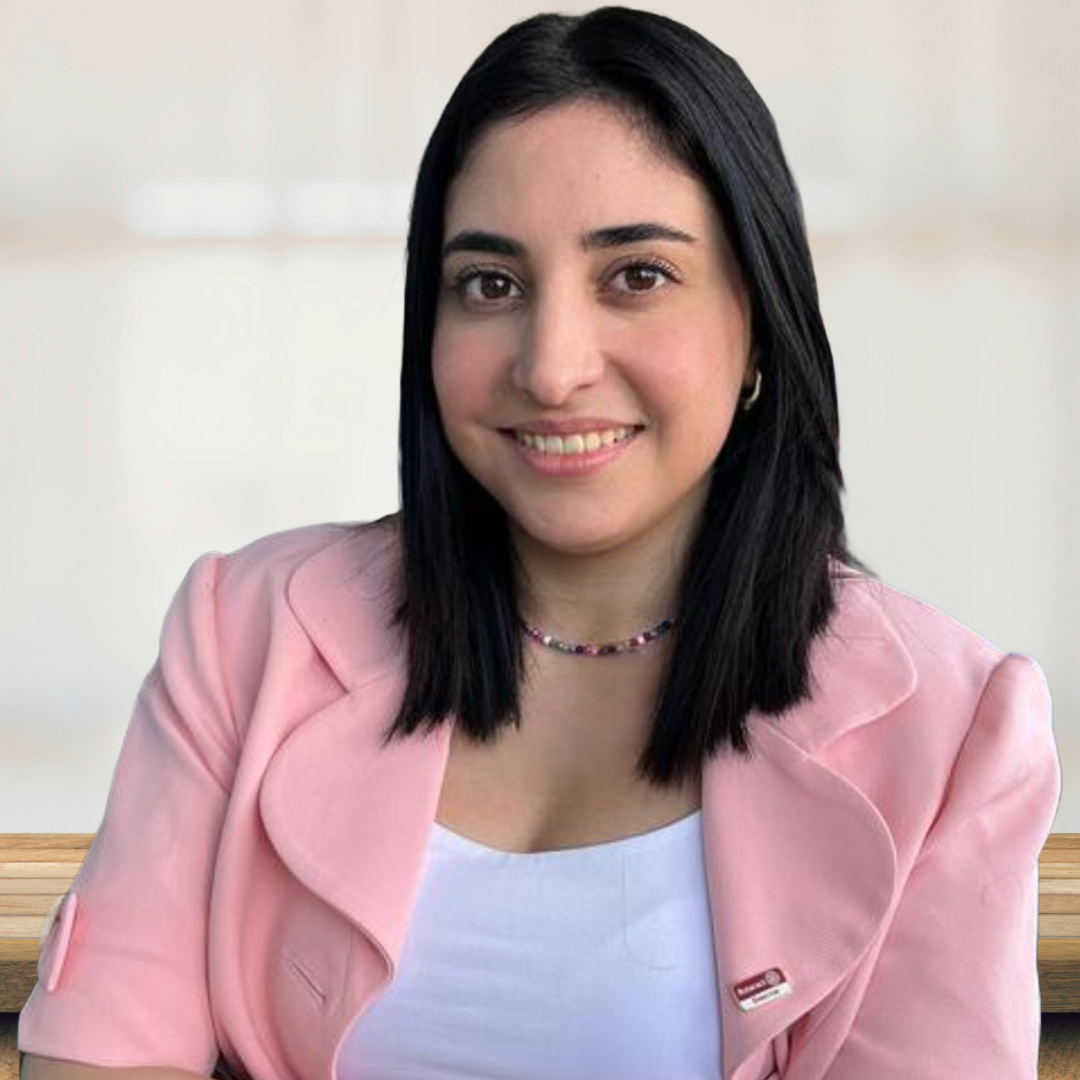 A woman with shoulder-length black hair, wearing a pink blazer over a white top, smiling at the camera, seated at a wooden table with a plain light-colored background.