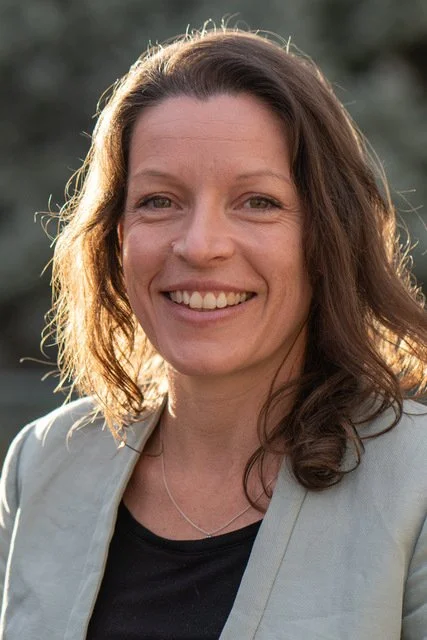 A woman with shoulder-length brown hair smiling outdoors sunlight behind her shoulders, wearing a light-colored blazer over a black top.