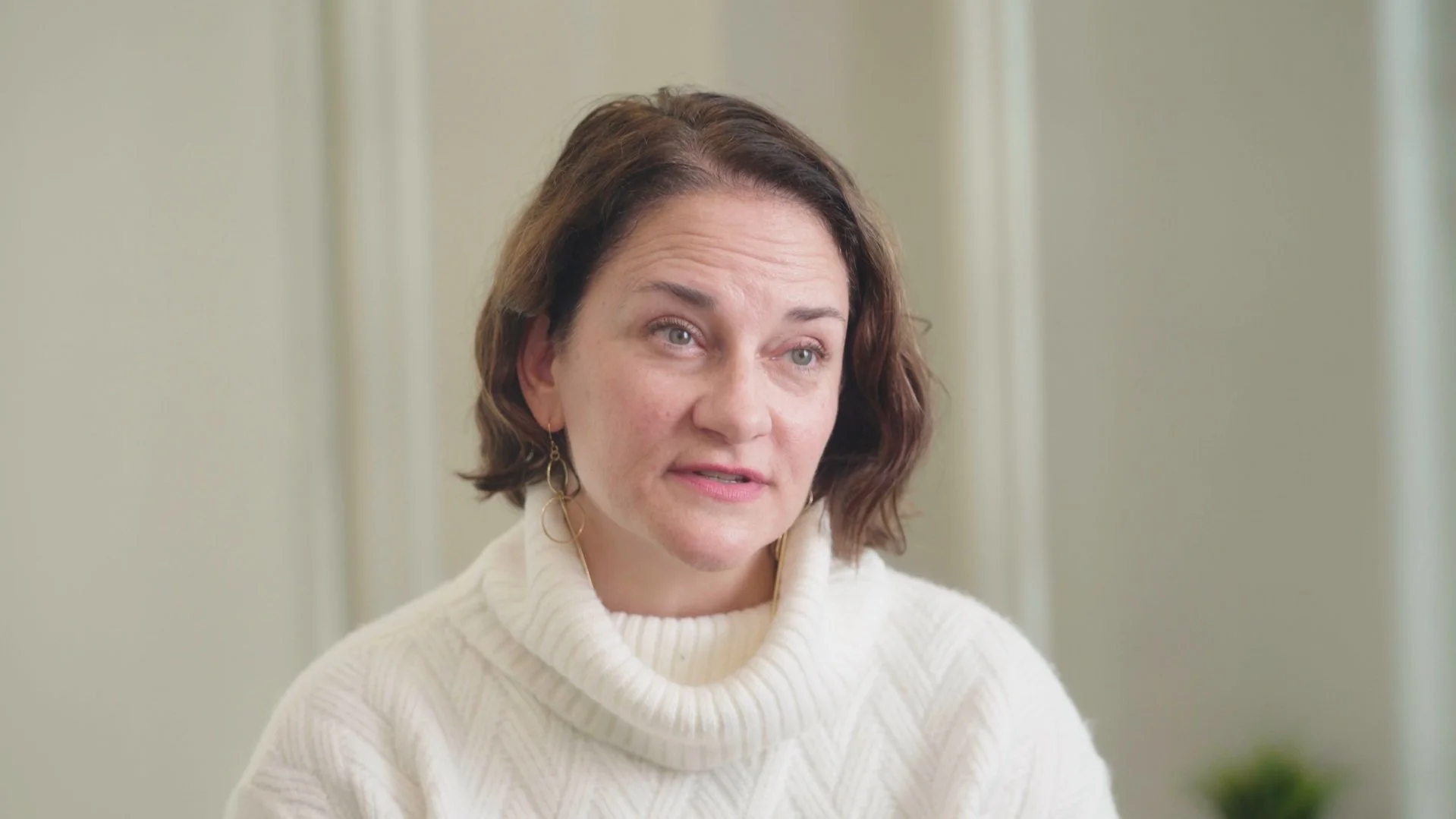A woman with short brown hair wearing a white turtleneck sweater and gold earrings, sitting indoors against a neutral background.