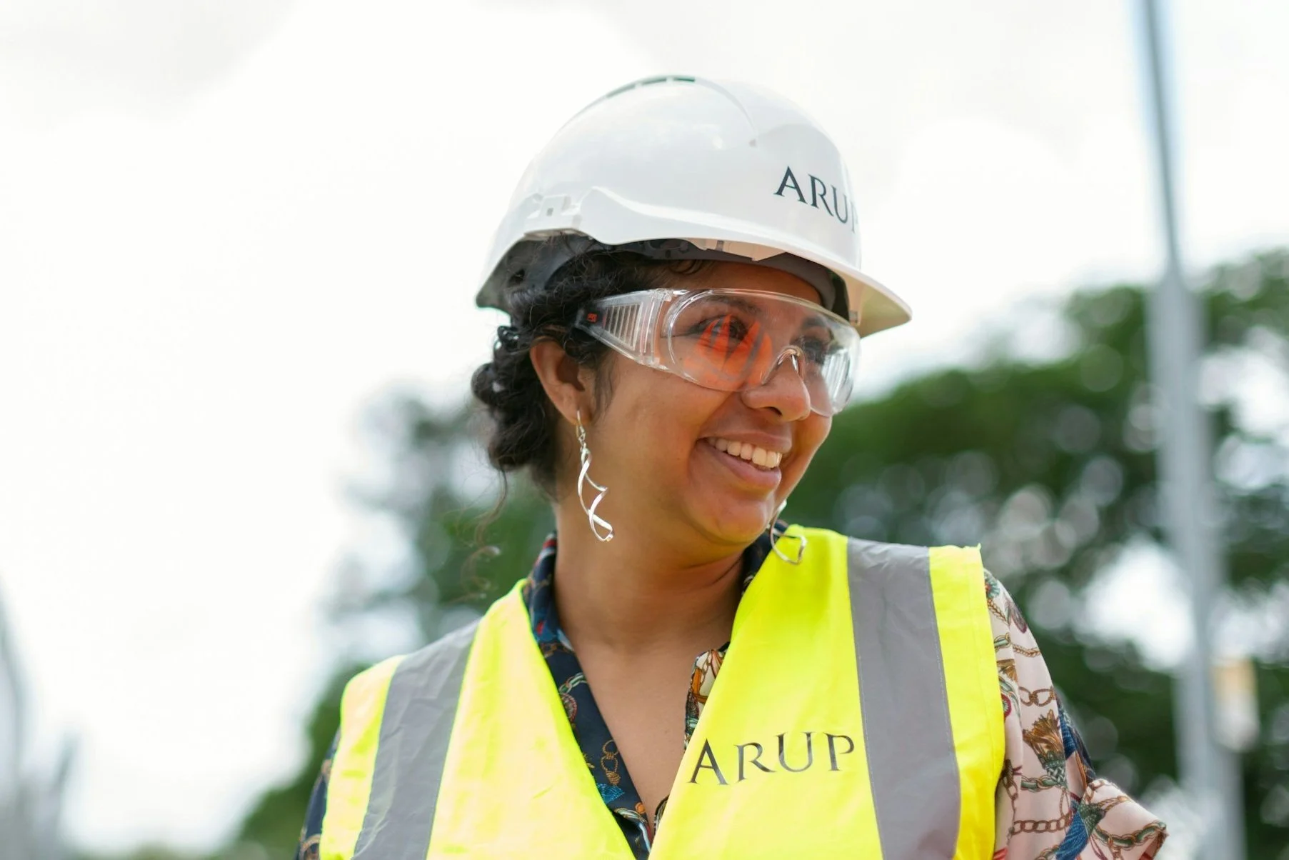 A woman wearing a white safety helmet, clear safety goggles, and a high-visibility yellow vest with 'ARUP' logo. She is smiling and looking to her left. She has dark curly hair and earrings. The background shows blurred green trees and outdoor environment.