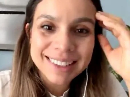 A woman smiling with long brown hair, wearing earphones, in a bright room.