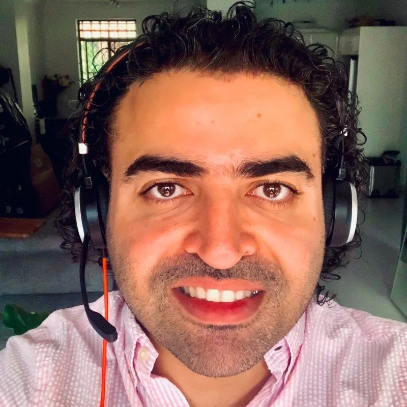 A man with curly dark hair wearing a pink checkered shirt and a headset, smiling at the camera indoors.