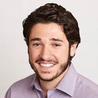 A smiling man with dark wavy hair and a beard, wearing a light purple shirt, posing against a plain background.