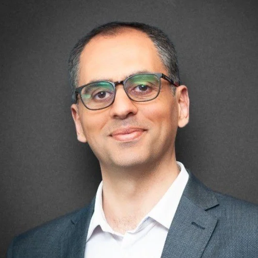 Headshot of a man wearing glasses, a white shirt, and a dark suit jacket, against a dark background.