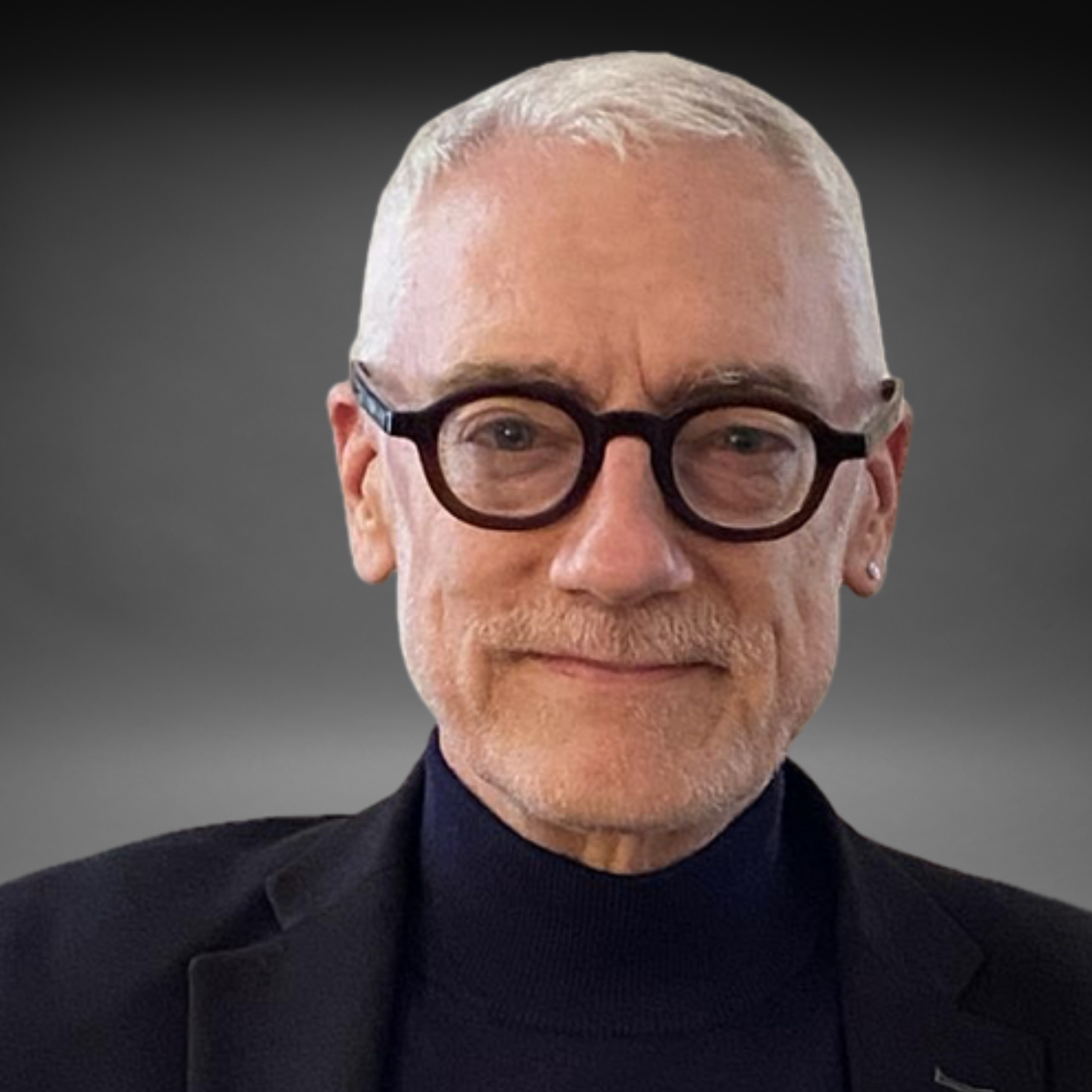 A close-up portrait of an older man with light skin, white hair, glasses, and a short white beard, wearing a black turtleneck and blazer against a dark background.
