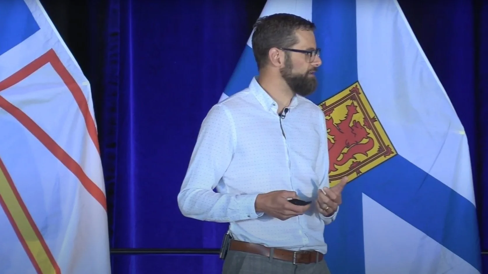 A man with glasses and a beard standing on stage holding a remote, with Newfoundland and Nova Scotia flags in the background.