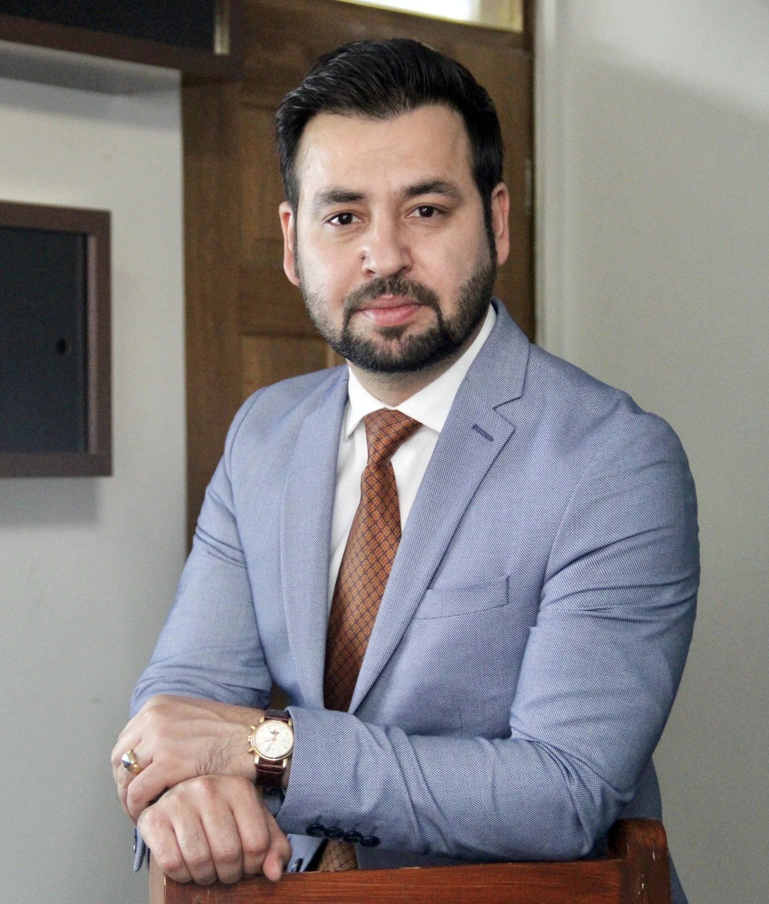 A man with dark hair, beard, and mustache wearing a light gray suit, white shirt, and brown patterned tie sits with arms crossed, looking at the camera, in an office setting.