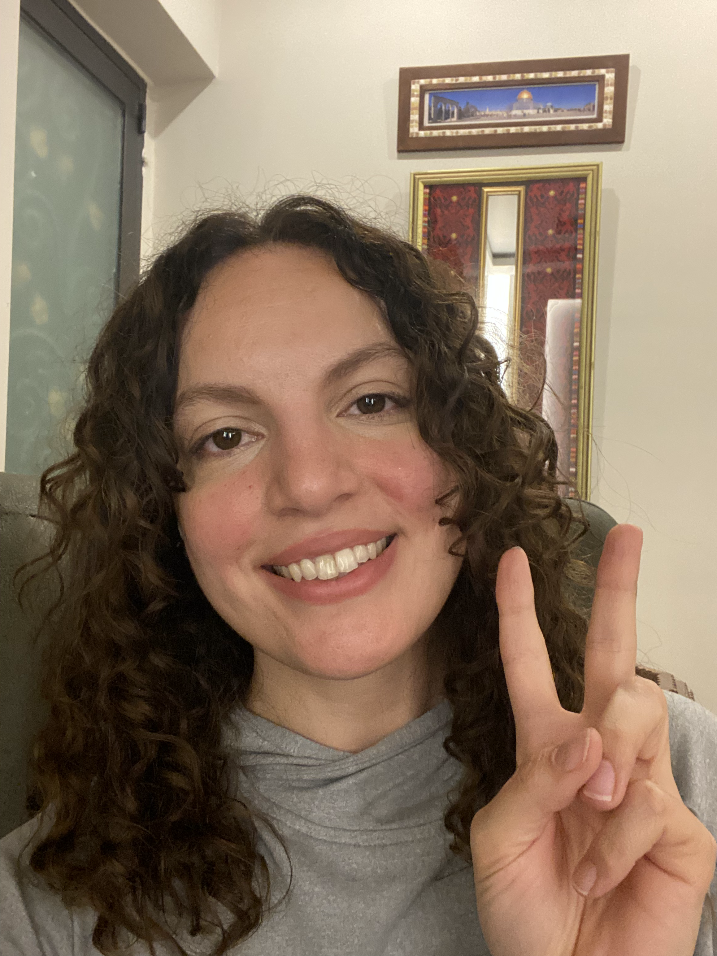 A woman with curly brown hair making a peace sign gesture and smiling, indoors with framed artwork in the background.