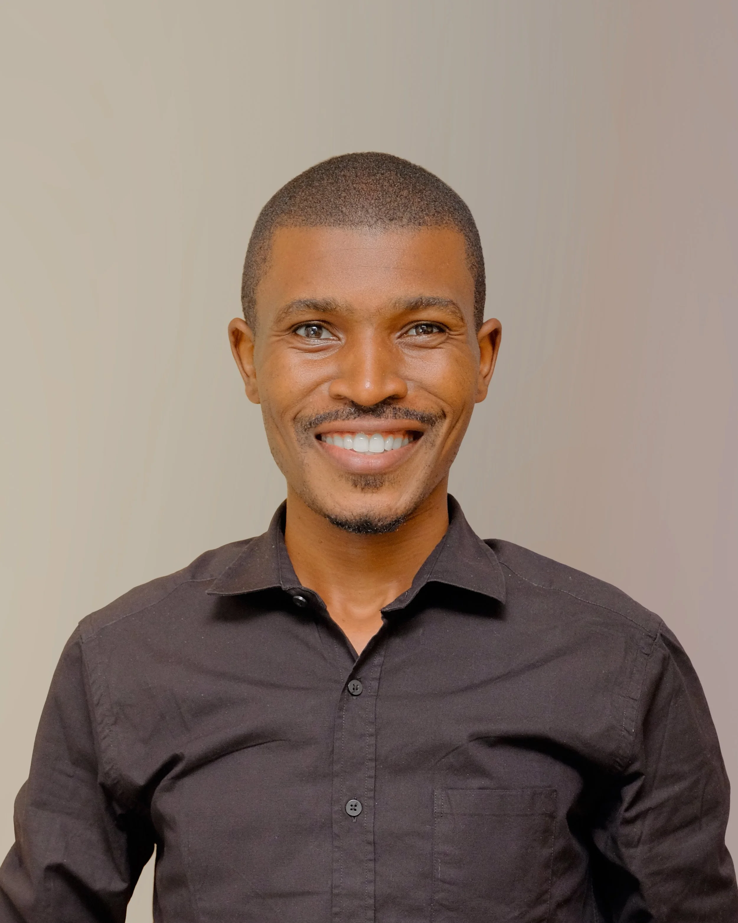 Headshot of a smiling man with short hair, a goatee, wearing a black button-up shirt, against a neutral background.