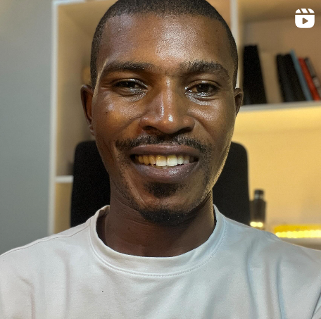 A smiling man with short black hair, a goatee, and wearing a white T-shirt, sitting in front of a bookshelf and a yellow-lit desk.