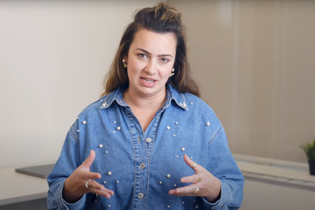 A woman with long brown hair wearing a blue denim shirt decorated with pearl-like beads, making expressive hand gestures while speaking in an indoor setting.