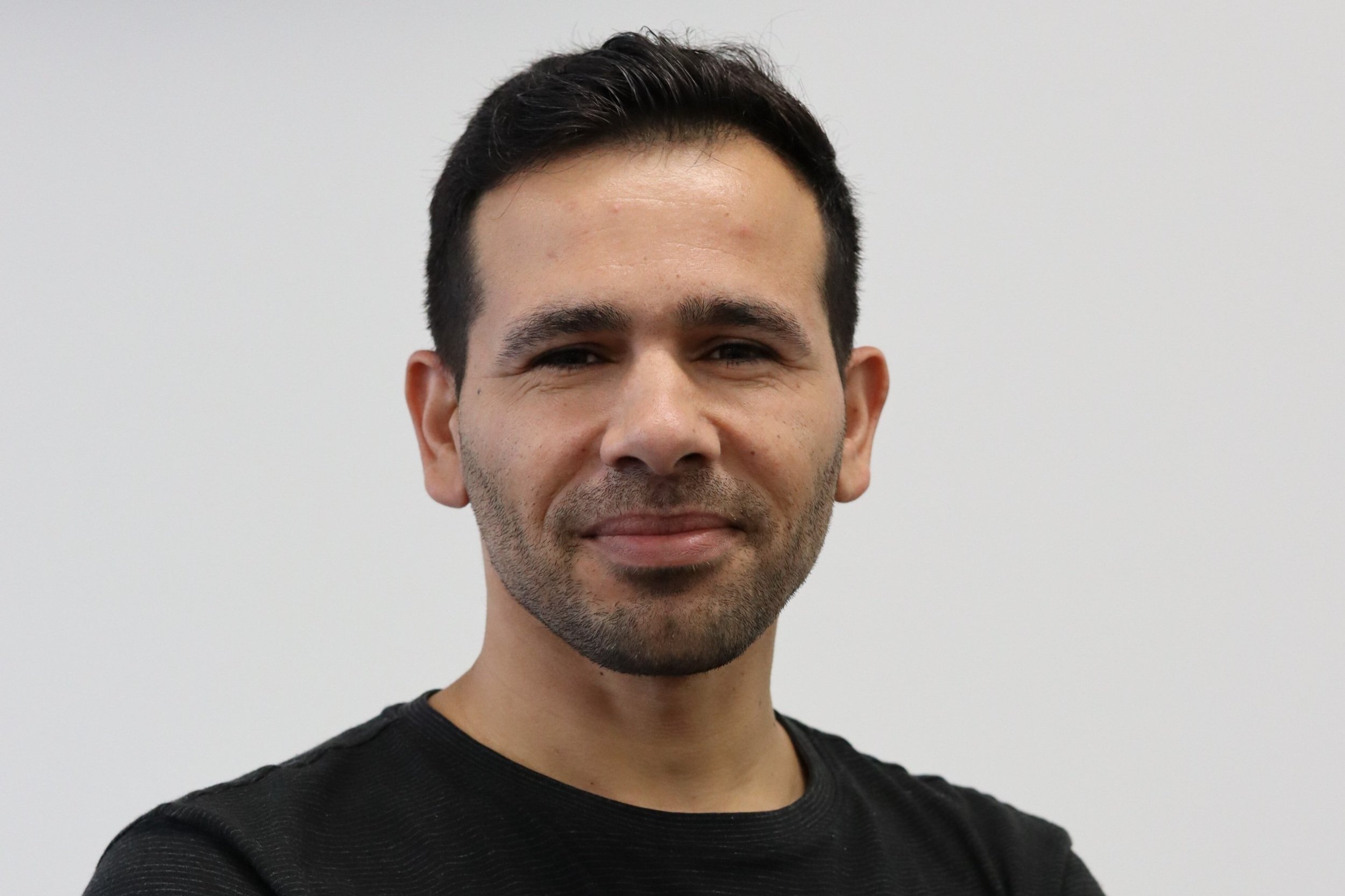A man with short dark hair, light skin, and slight beard, wearing a black shirt, standing against a plain white background.