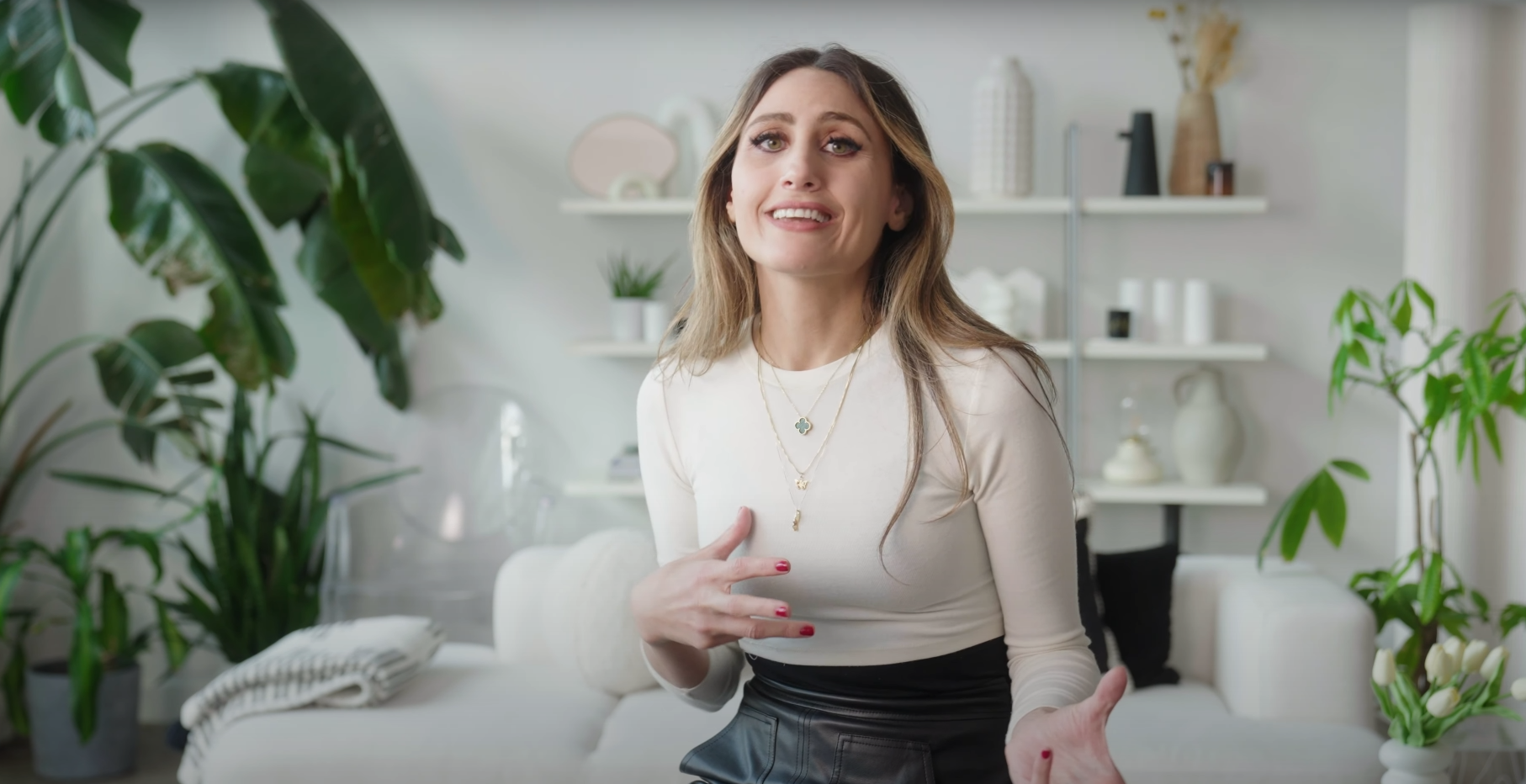 A woman with long light brown hair, wearing a white long-sleeve top and layered necklaces, appears to be speaking or explaining something in a bright, modern living room with green plants and white decor.