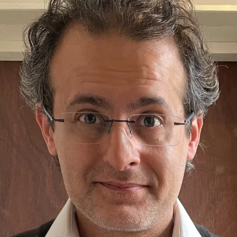 A middle-aged man with curly salt-and-pepper hair, wearing glasses and a collared shirt, is smiling at the camera in an indoor setting.