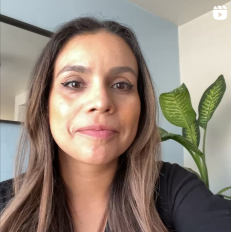 A woman with long hair and makeup taking a selfie in front of a plant with large green leaves.