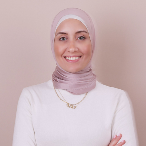 Portrait of a woman wearing a light pink hijab, white top, and gold necklace, smiling at the camera.