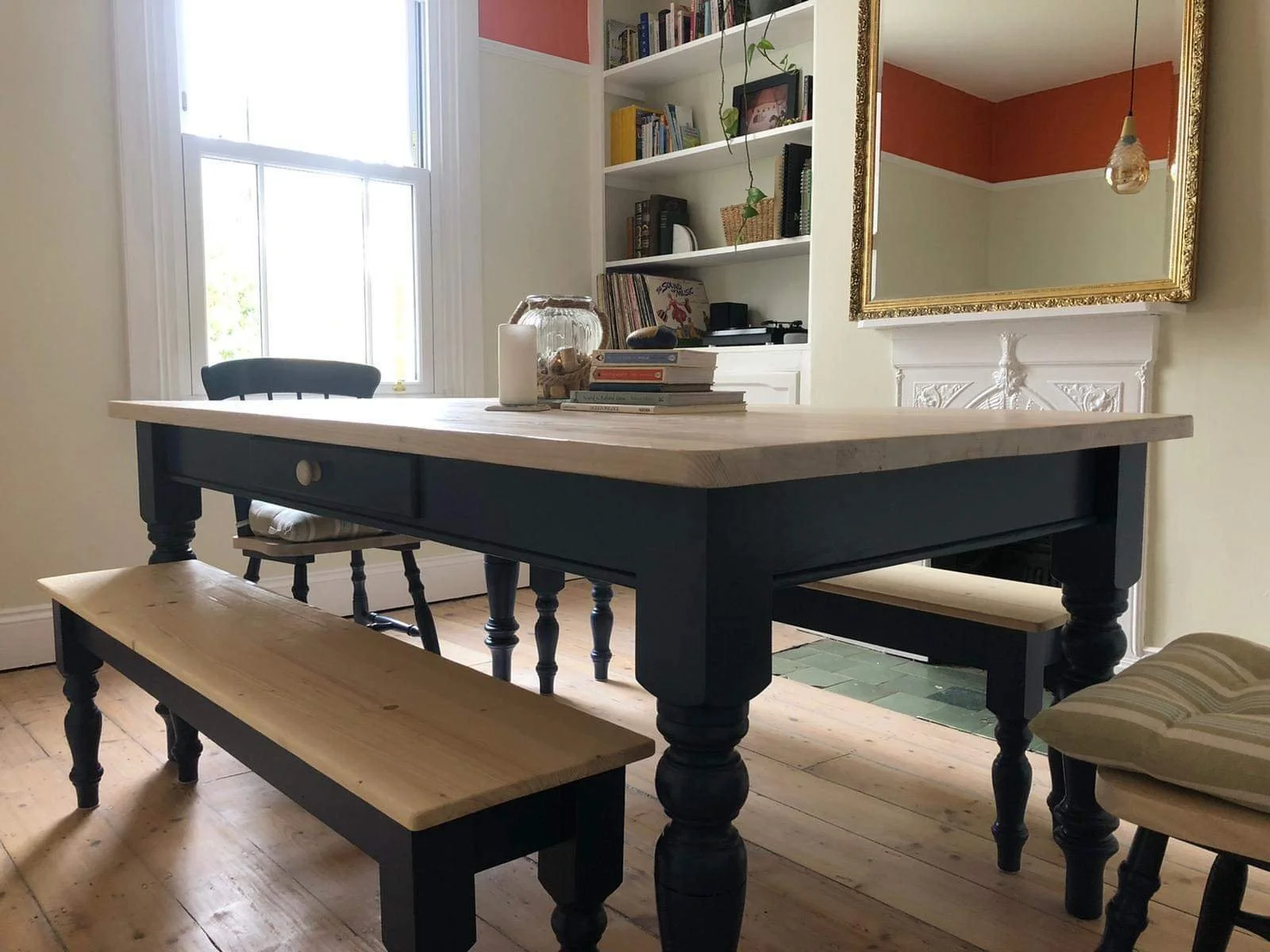 Dining room interior with a stiffkey blue painted wooden dining table with light wooden top, matching benches, and a cushioned chair. The room features a large window, built-in shelves with books and décor, a decorative mirror, a pendant light.