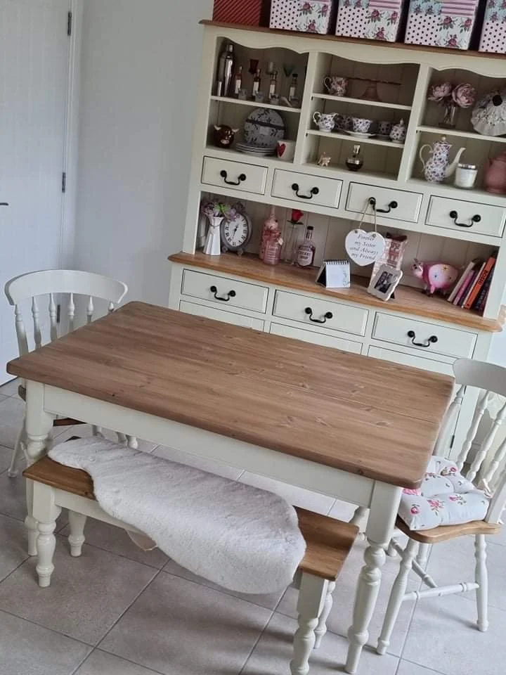 Vintage-style dining area with a cream painted farmhouse table and chairs, complemented by a white hutch displaying decorative china and items, including floral-patterned teapots and boxes, with a cozy sheepskin throw on the bench.