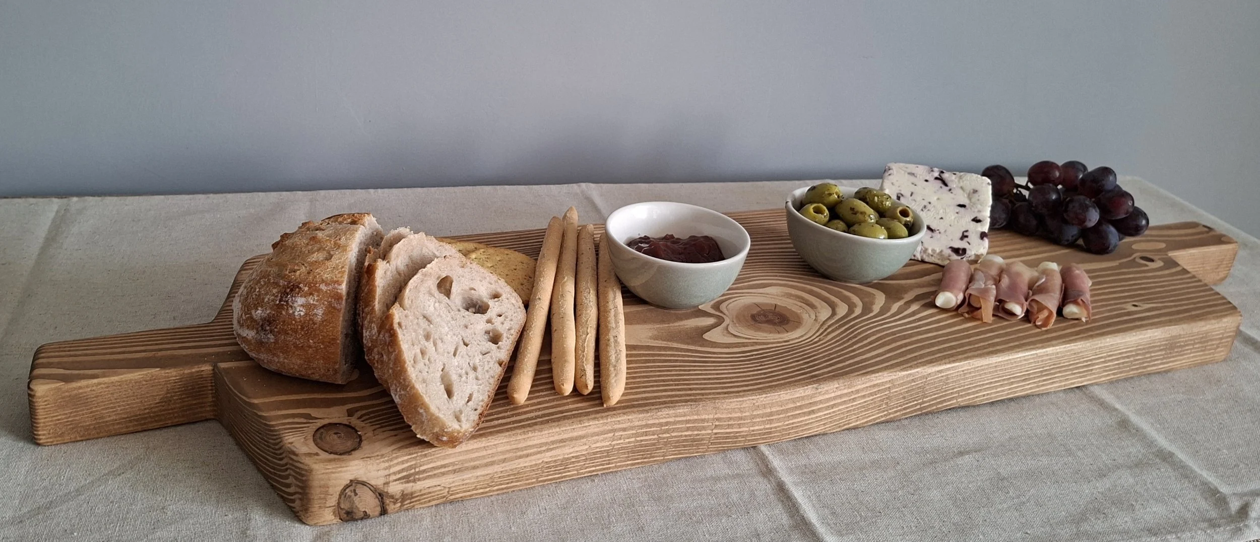 A wooden serving board with bread, breadsticks, jam, green olives, blue cheese, grapes, and rolled ham and cheese slices.