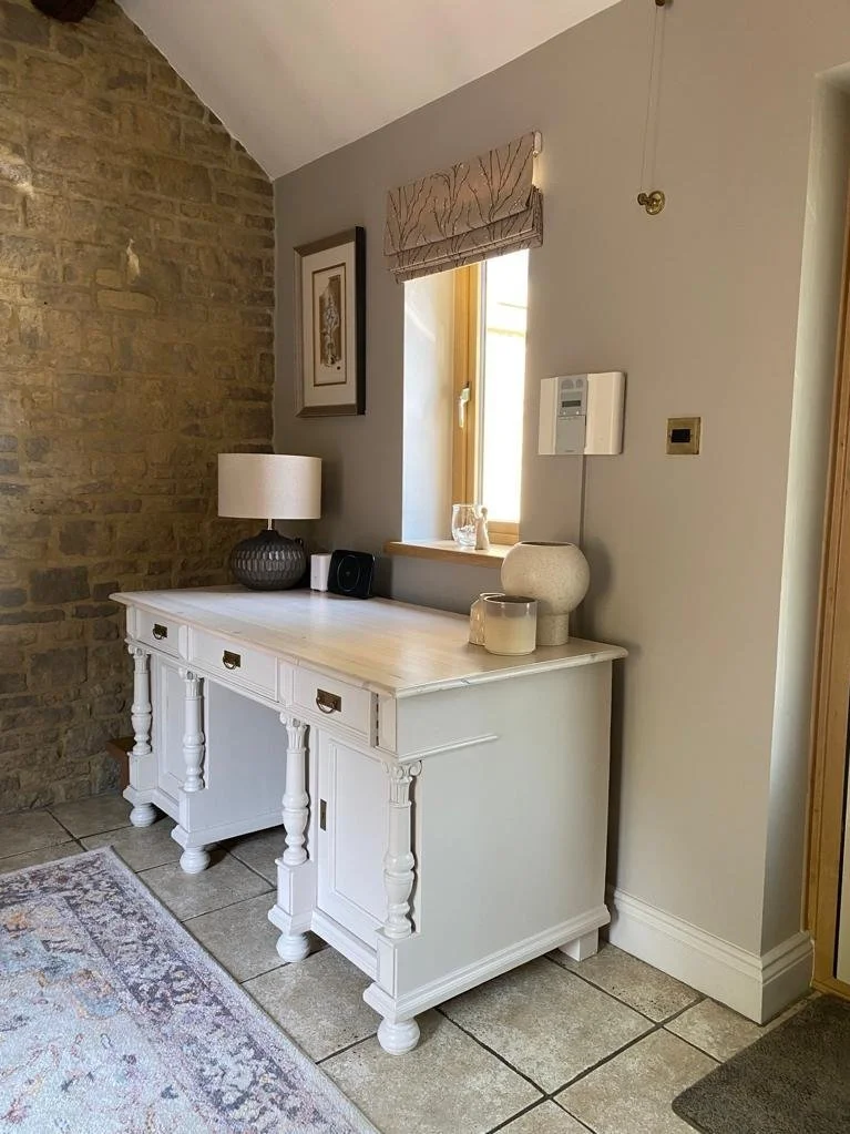 A white vintage-style sideboard with drawers and cabinet doors, placed against a wall near a window. The sideboard has various decorative items on top, including a table lamp, a black speaker, ceramic vases, and candles. The room features a brick acc