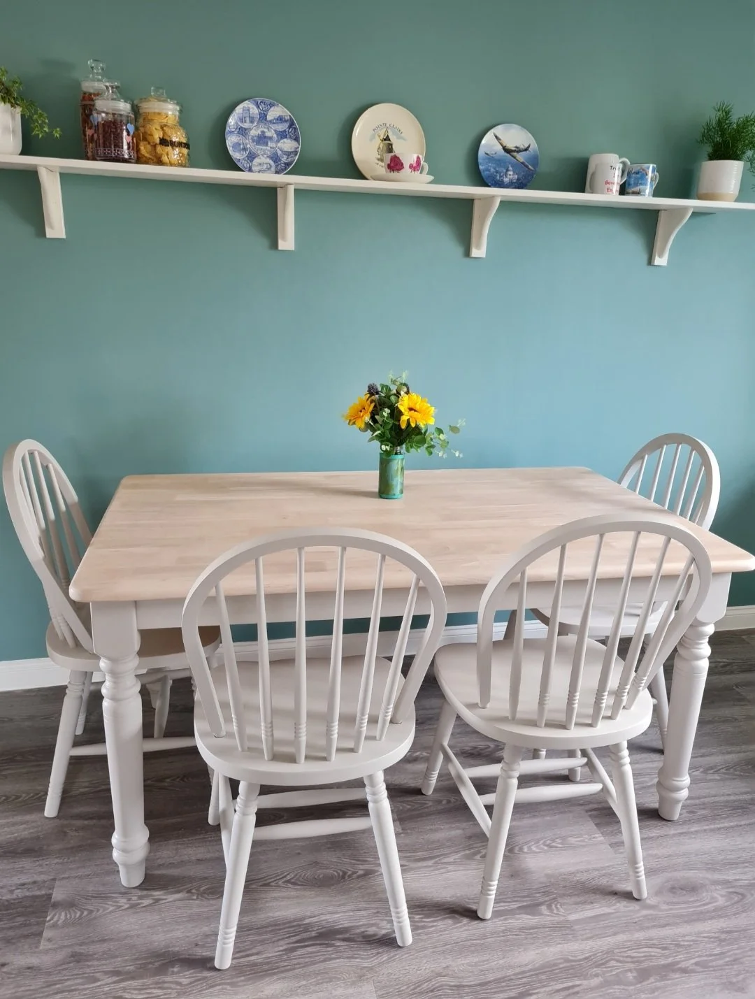 Neutral painted and whitewashed light wooden dining table with four matching painted chairs and a vase with sunflowers, against a green wall with a shelf displaying decorative plates, jars, and plants.