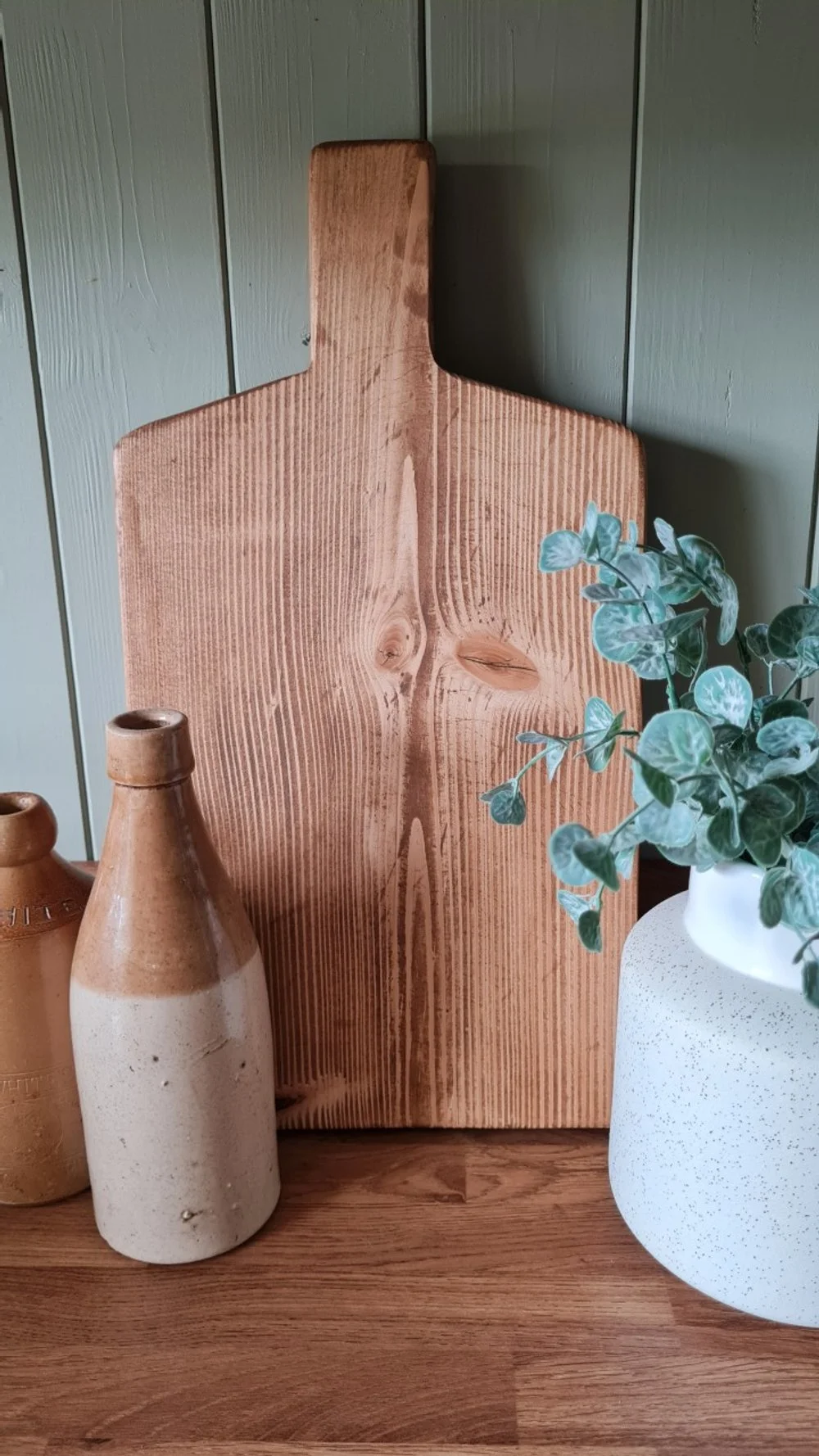 A wooden cutting board with a handle, placed against a greenish wall. Two ceramic bottles and a potted plant are on a wooden surface in front of it.