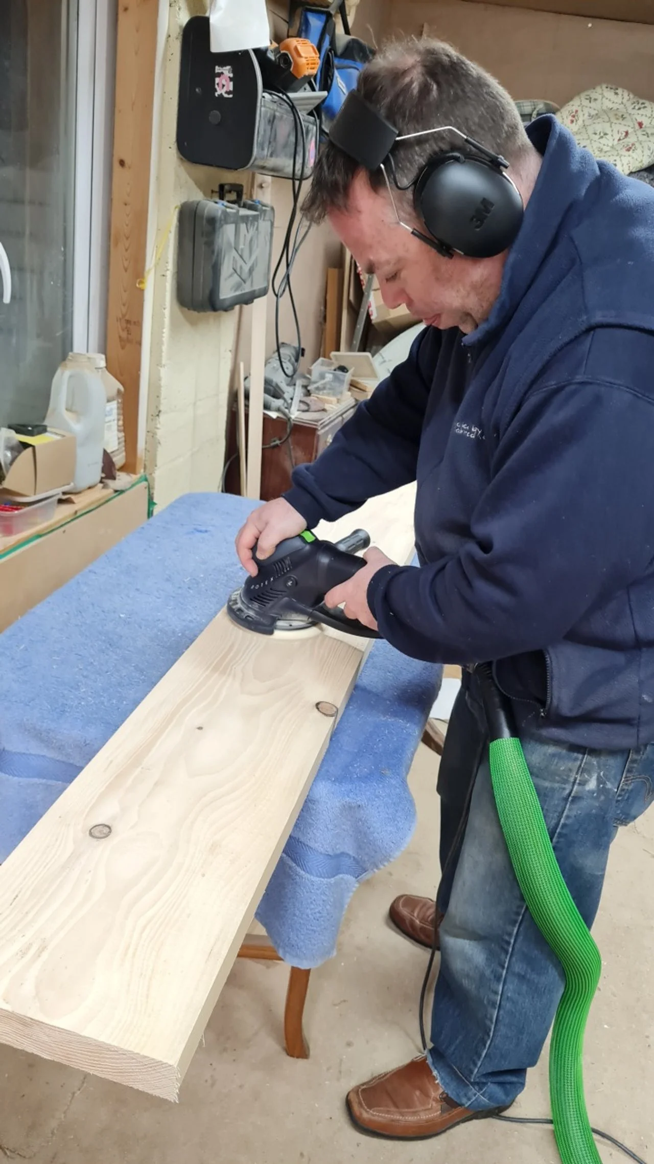 A man with hearing protection and glove uses a power sander on a wooden board in a workshop.