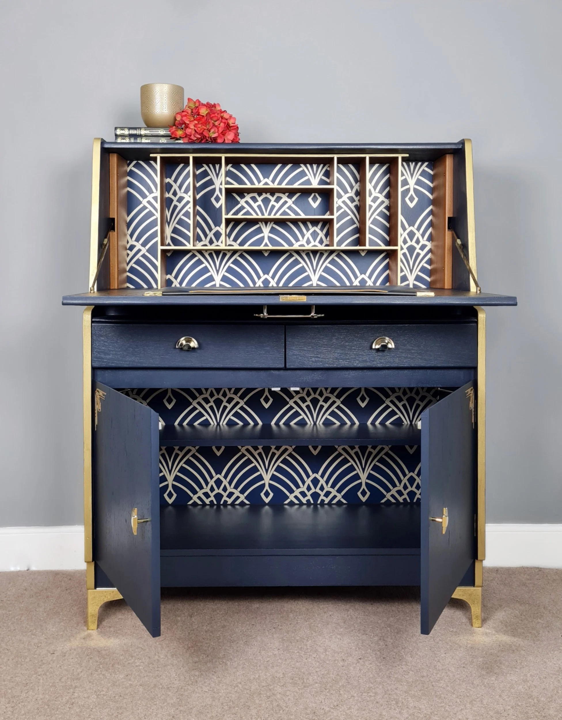 A navy blue and gold vintage secretary desk with open lower cabinet, two drawers, and intricate art deco patterned wallpaper inside.