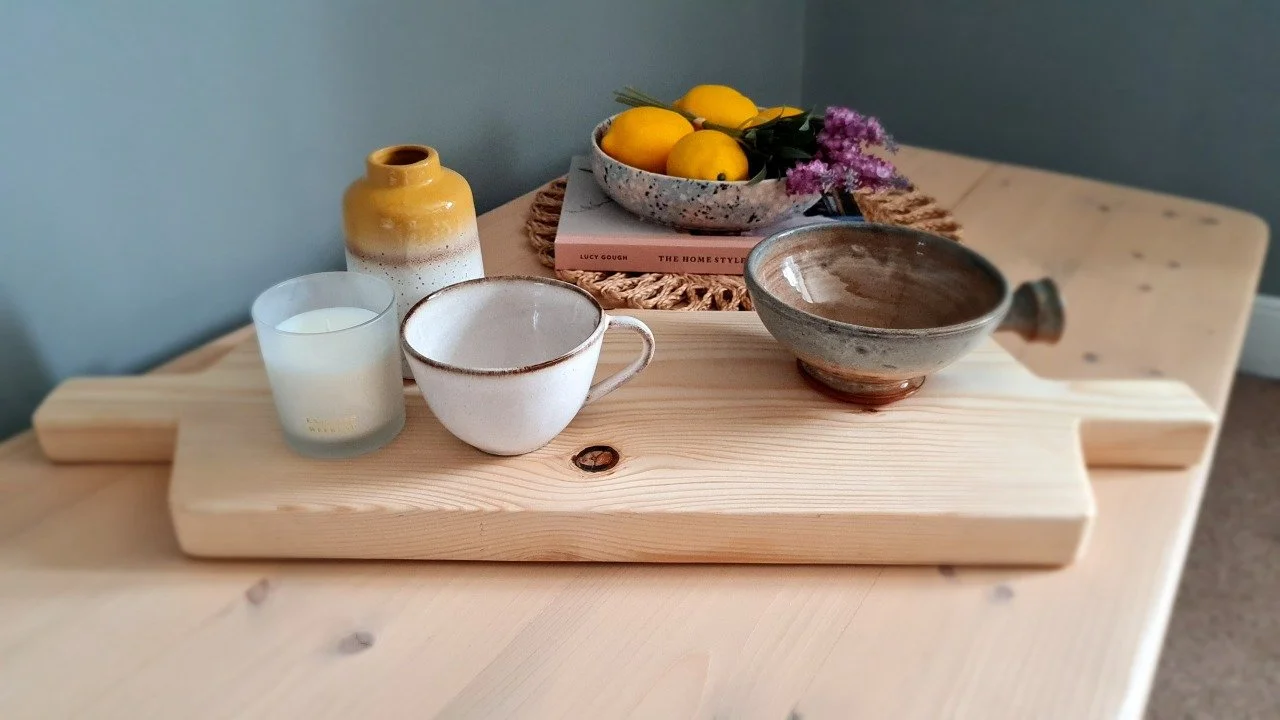 Decorative wooden tray with a yellow and white ceramic vase, a glass candle holder with a white candle, a white mug with a handle, a bowl with a spout, and a bowl of yellow lemons with purple flowers and a book underneath.