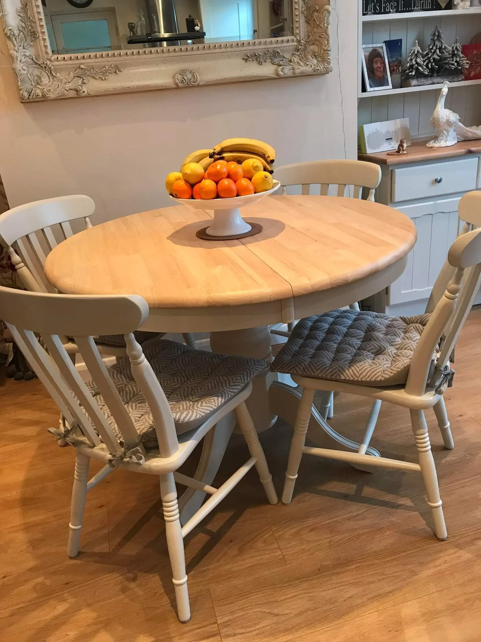Round painted extendable wooden dining table with cushioned chairs, a fruit bowl containing bananas, oranges, and lemons, and a wall mirror in the background, set in a cozy dining area with decorative items and shelving.