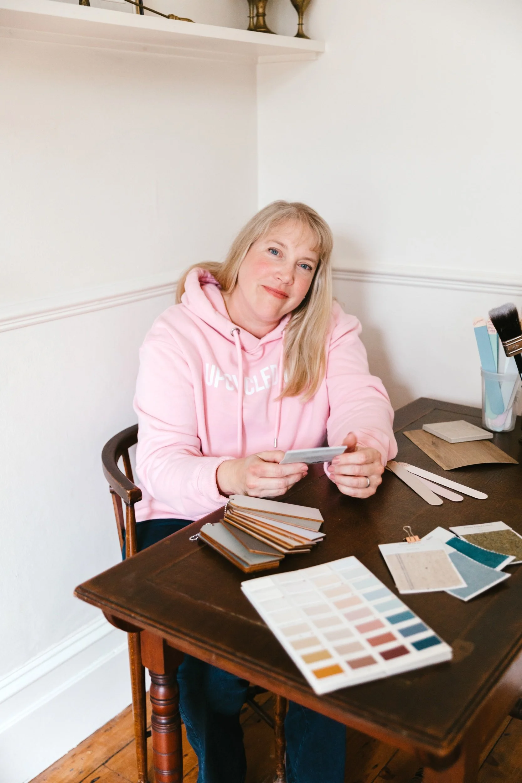 Woman sitting at a wooden table with color swatches, paint samples, and design materials, wearing a pink hoodie.