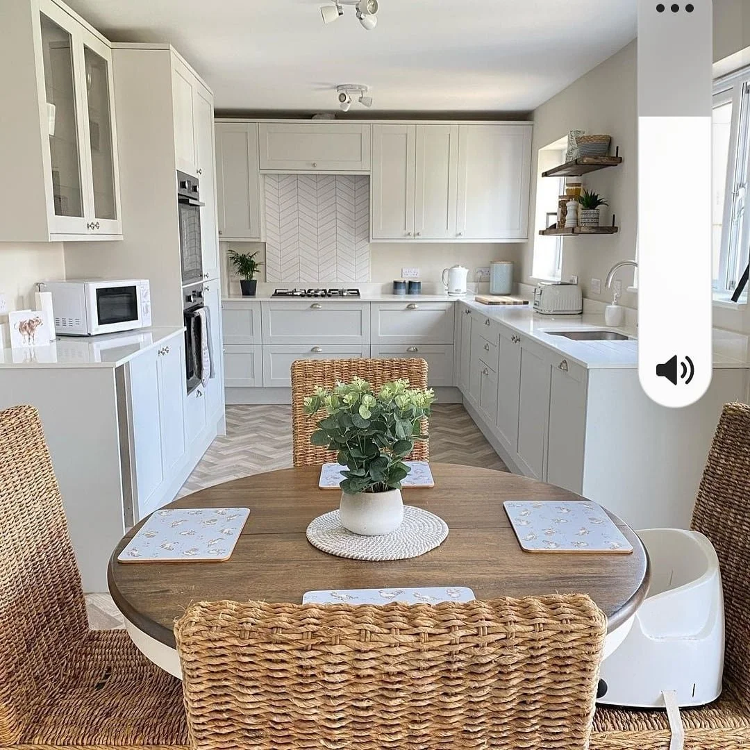 Modern kitchen and dining area with white cabinets, wicker chairs, and a round painted wooden table with walnut stained top and a plant. Features include a stove, microwave, and white countertops.