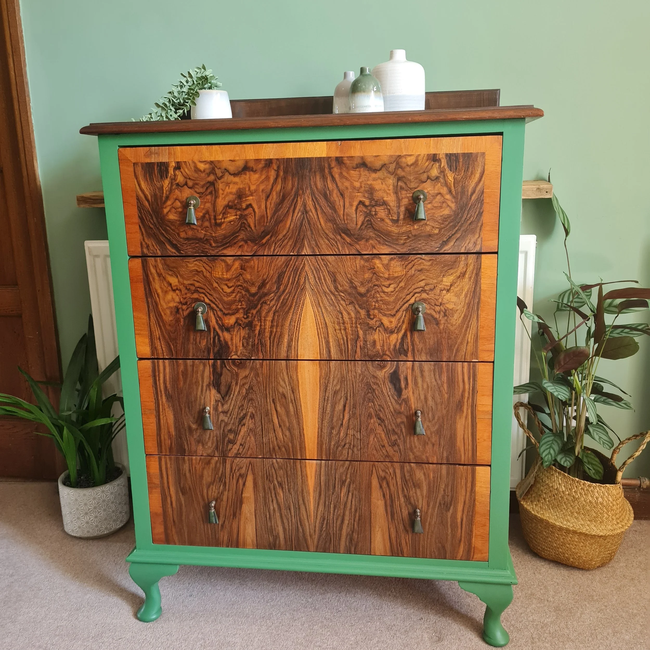 A vintage wooden dresser with natural wood grain front, green painted sides and legs, and five drawers with small metal knobs. On top, there are three decorative vases and a small white planter with a green plant. Two potted plants are beside the dre
