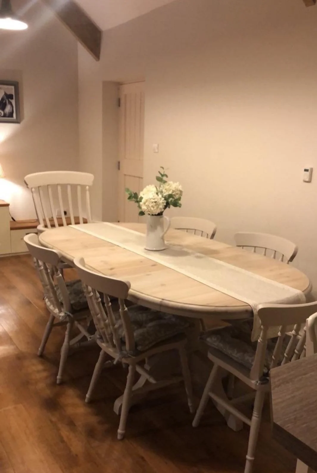 Large oval extendable painted taupe wooden table and six matching painted chairs set in a Dining Room, featuring a vase with white flowers on the table.