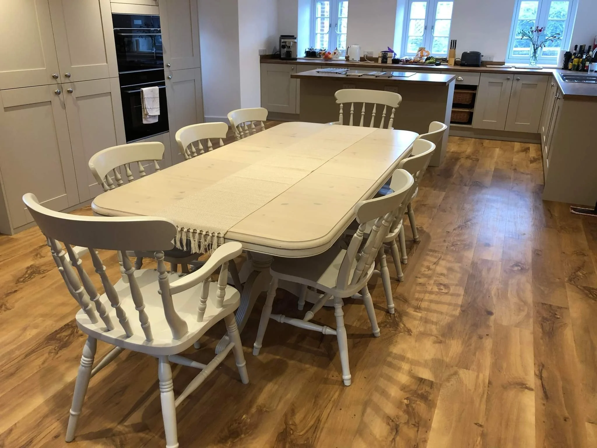 Kitchen with a taupe painted and whitewashed wooden dining table and eight matching painted chairs, wooden floor, pale cabinets, and appliances such as an oven and toaster.