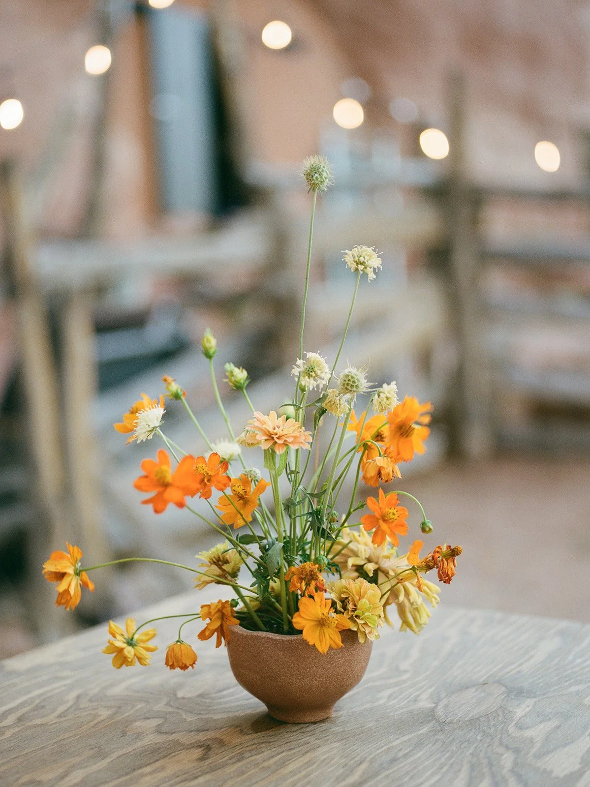 Farm Yard - Moab Utah - A brown ceramic vase with orange and cream flowers on a wooden table, with blurred string lights and shelves in the background.