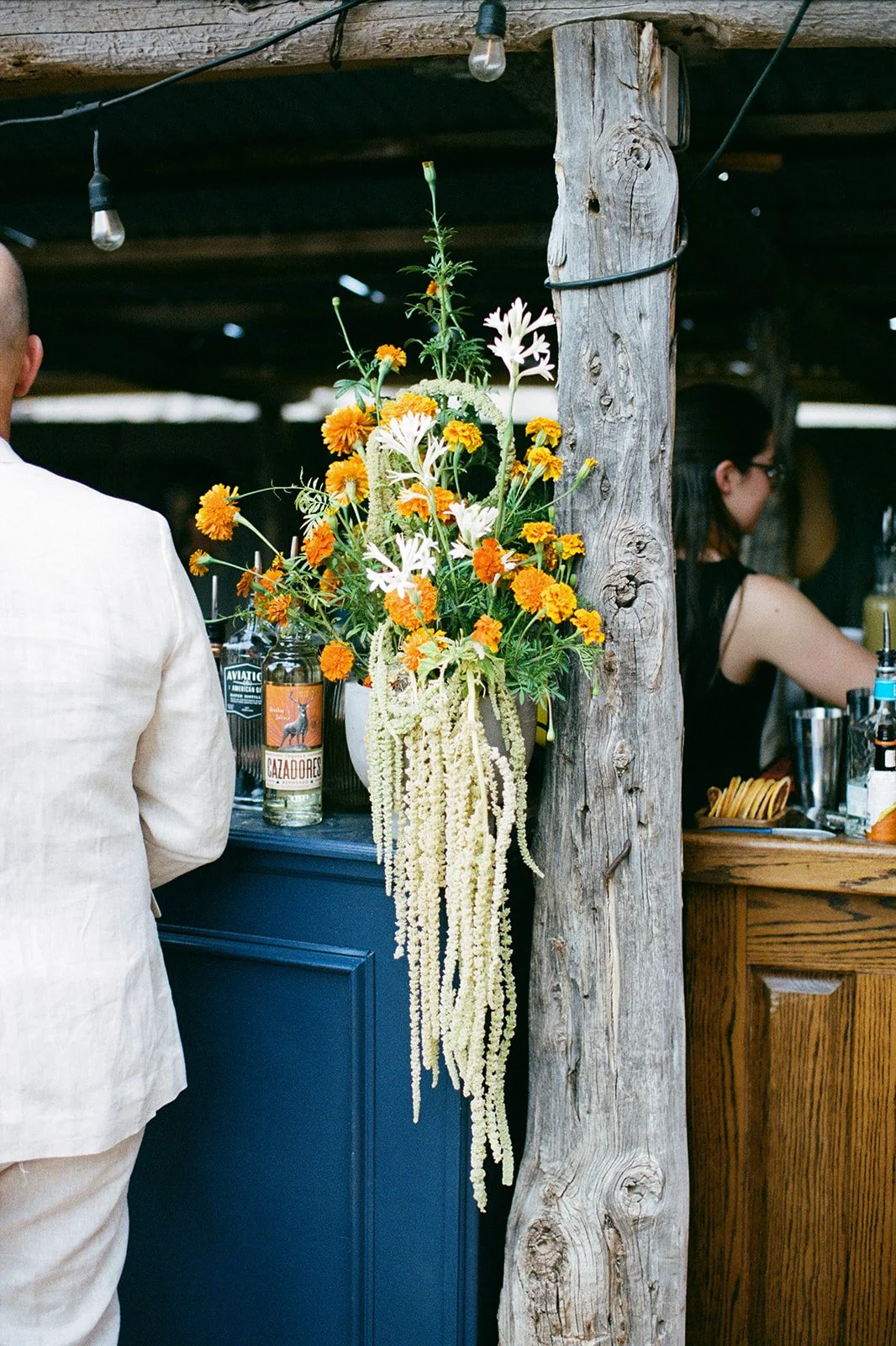 Farm Yard - Moab Utah - Colorful flower arrangement with orange, white, and green flowers hanging on a rustic wooden post at a bar or outdoor venue.