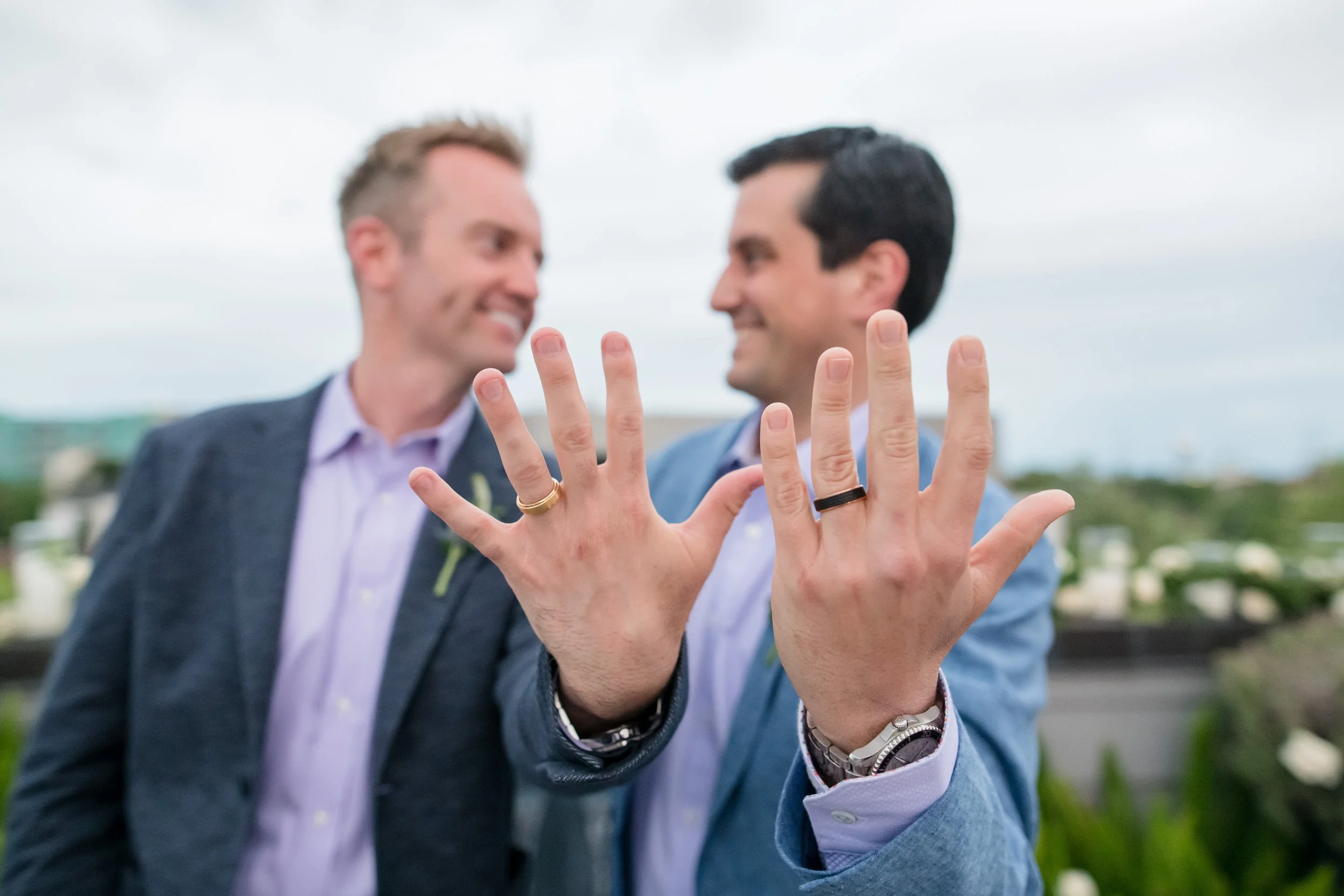 Two newlywed grooms smiling behind their raised hands as they show their wedding rings.