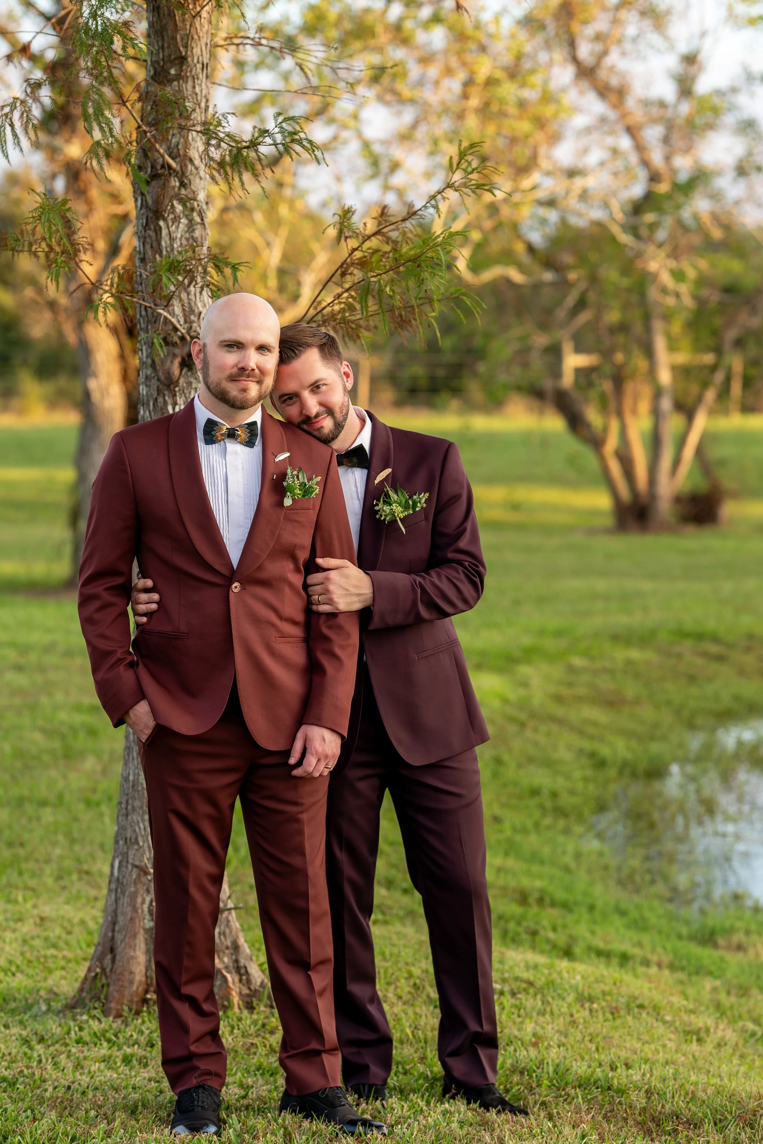 Two grooms in coordinating burgundy suits pose together beside a tree and pond.