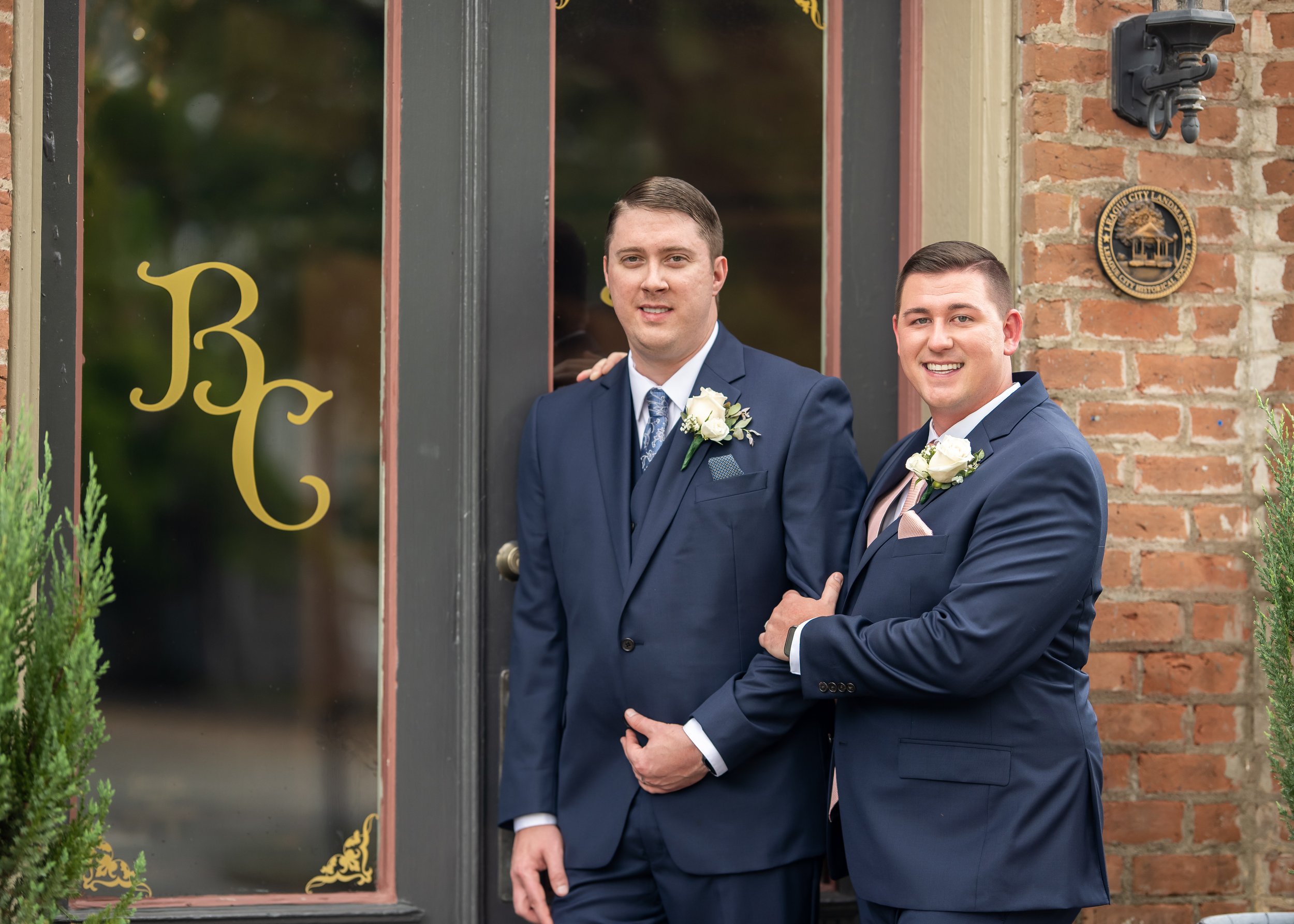 Two grooms in navy suits pose outside Butler’s Courtyard in League City, Texas.